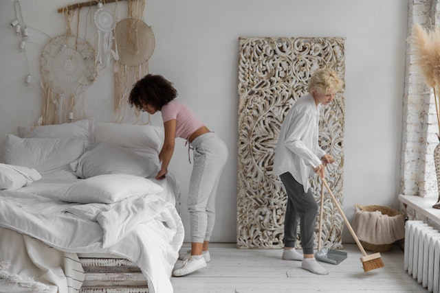 Two people cleaning a bedroom, one is sweeping and the other is making the bed